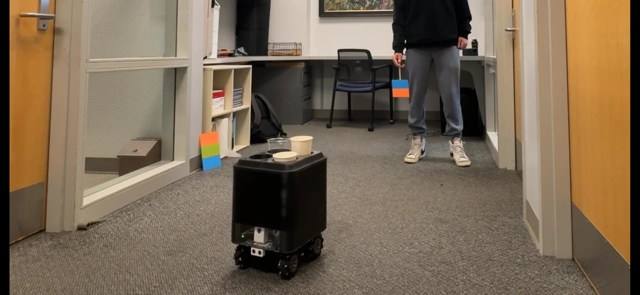 Black four-wheel delivery robot carrying cups through a doorway, with its front camera and ultrasonic sensor visible.